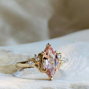 Gold engagement ring with light pink long marquise shaped center and delicate floral elements on the sides made of carved gold and small diamonds