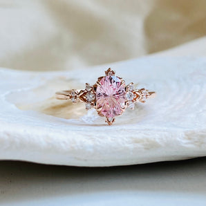 Gold engagement ring on white background. Center clear light pink oval gem has a carved star design inside the stone. It is surrounded by delicate clear diamonds and gold beads, that create unique fantasy designs. The top and bottom of the center stone has a tiny gold and diamond tiara, fit for a princess 